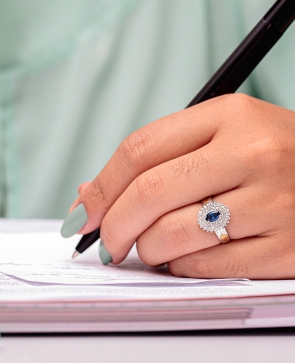 Mulher escrevendo em um papel, ela usa anel de formatura de prata com pedra azul e várias pedras brancas ao redor