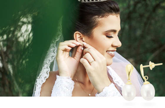 Noiva com vestido de casamento colocando brinco de ouro e pérola, ela usa também anel de noivado e colar com pérola. 