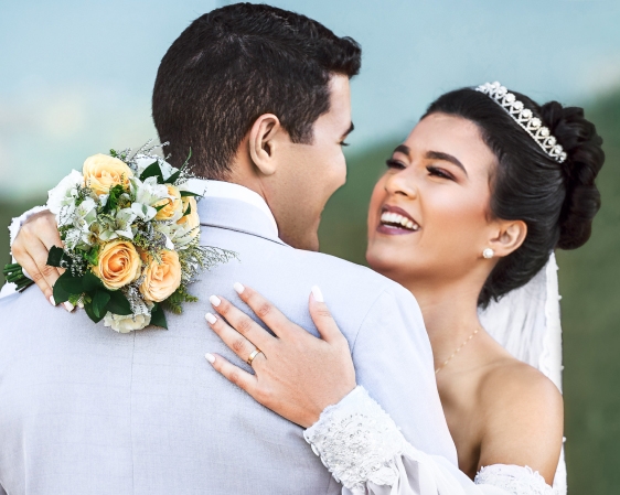 Noivos em um campo, abraçados e sorrindo. A noiva segura o buquê e na mão esquerda usa uma aliança de casamento.