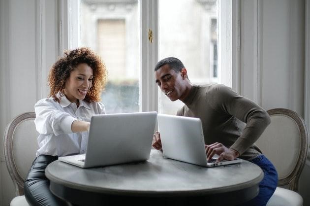 Casal em casa, cada um com seu computador, a mulher está mostrando algo no seu computador para o namorados