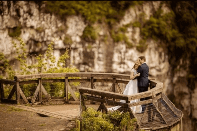 Homem e mulher com trajes de casamento em uma alta estrutura de madeira em Gramado. Ao fundo tem um paredão de pedras. 