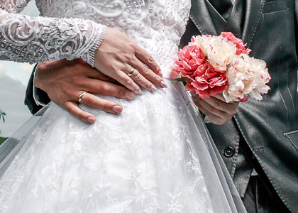 Noivos em trajes de casamento, o noivo abraça a cintura da noiva, eles usam alianças de casamento tradicionais em ouro 18k.