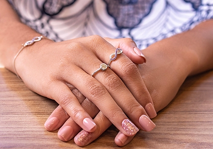 Mãos de uma mulher sobrepostas na mesa. Ela usa um mix de anéis: um com o símbolo do infinito e um anel chuveiro. 