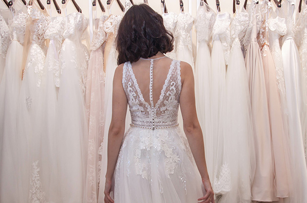noiva de costas com vestido branco de casamento olhando uma arara de vestidos de noivas, todos de cor branca e off-white. 