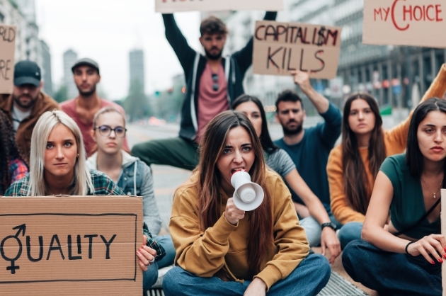 Mulheres sentadas em protesto  com placas pedindo igualdade, ao centro da imagem mulher com megafone