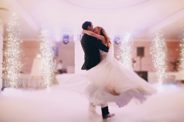 Salão de festa de casamento fogos saem do chão enquanto o noivo gira a noiva que enlaça seu pescoço em um abraço
