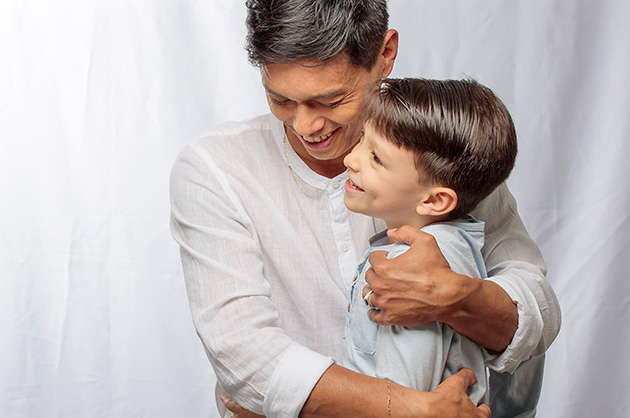 pai e filho abraçados, pai de camisa branca e filho de camisa azul claro, ambos sorrindo 