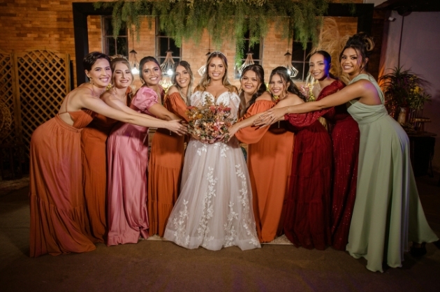Noiva sorridente segura buquê a frente de seu corpo, as madrinahs estendem a mão para tocar as flores e sorriem para a câmera
