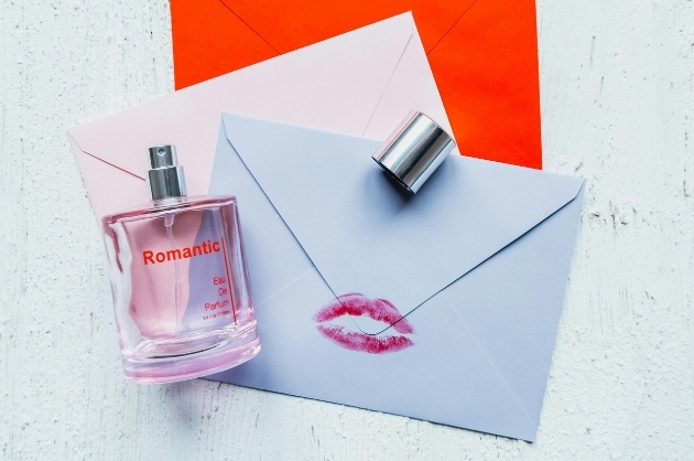 Envelope azul claro tem como o selo a marca de batom de um beijo, em cima um frasco de perfume