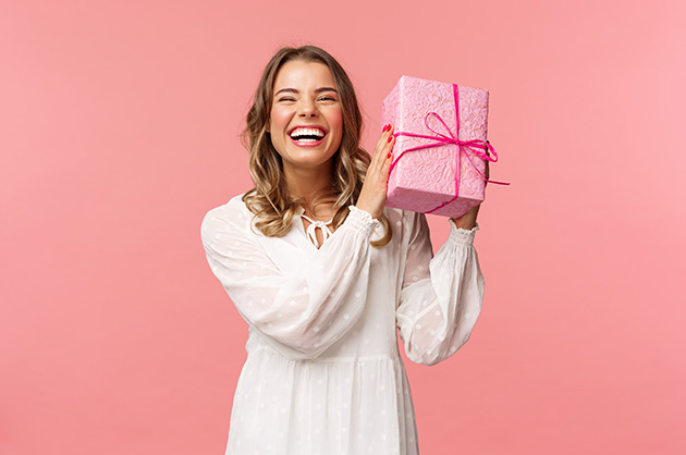 banner com fundo rosa com mulher loira de blusa branca segurando um pacote de presente de embrulho rosa claro e laço rosa escuro