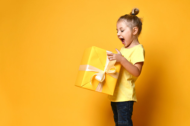 banner com fundo amarelo e criança de lado olhando muito feliz para o pacote grande de presente de cor amarela em suas mãos 